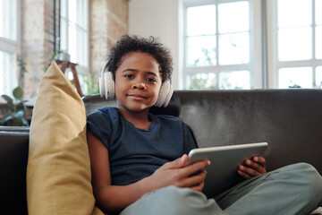 Happy little boy with headphones and tablet looking at you while watching online video