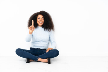 Young african american woman sitting on the floor showing and lifting a finger