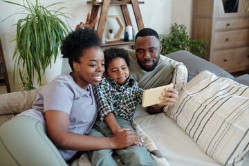 Joyful young African couple and their little son relaxing together on couch or bed and making selfie