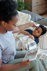 Young African female measuring body temperature of her sick little son lying in bed under blanket