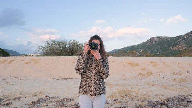 Front view smiling young casual woman on the beach holding camera and taking photo of beautiful landscape.