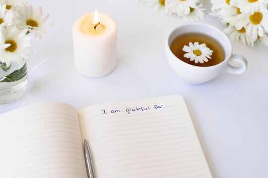 Close up of handwritten text "I am grateful for..." in foreground with notebook, pen, cup of tea, flowers