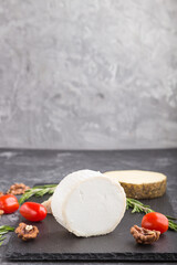 Goat cheese and various types of cheese with rosemary and tomatoes on black slate board on a black concrete background. Side view, copy space.