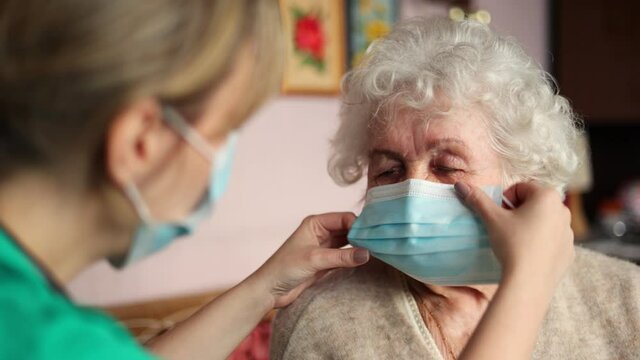 Female nurse helping a senior woman to put on protective face mask during home visit
