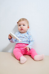 Fototapeta premium A little girl, eight months old, is sitting on the couch holding an ear piercing device. Beauty for young children