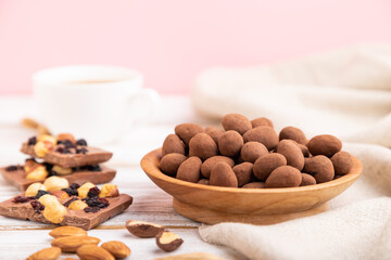 Almond in chocolate dragees in wooden plate and a cup of coffee on white and pink background. Side view, selective focus, copy space.