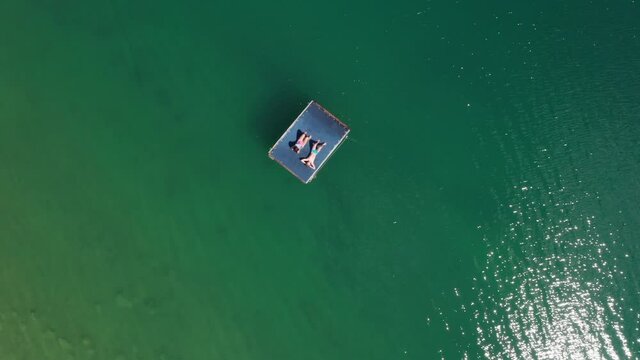 4K Sunbathing on turquoise lake floating platform mother and son aerial high altitude drone top view. Bright sun reflecting in green lake waves. Peruca Lake, Dalmatia, Croatia