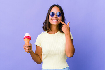 Young mixed race woman eating an ice cream smiles, pointing fingers at mouth.