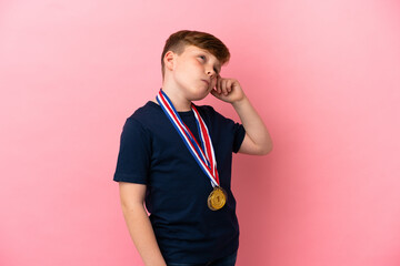 Little redhead boy with medals isolated on pink background having doubts and with confuse face...