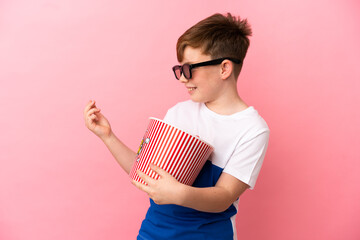 Little redhead boy isolated on pink background with 3d glasses and holding a big bucket of popcorns while looking side