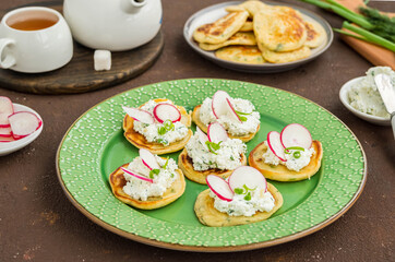 Snack mini pancakes with a spread of salted cottage cheese, herbs and slices of radish on a green plate on a brown concrete background.