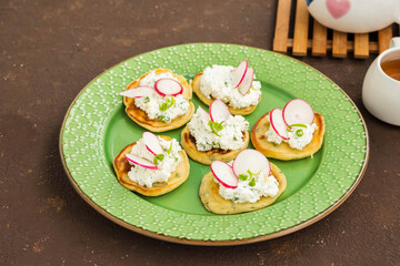 Snack mini pancakes with a spread of salted cottage cheese, herbs and slices of radish on a green plate on a brown concrete background.