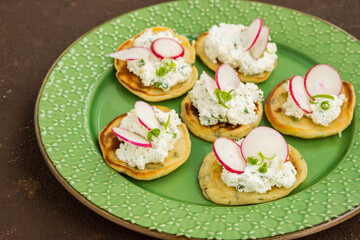Snack mini pancakes with a spread of salted cottage cheese, herbs and slices of radish on a green plate on a brown concrete background.