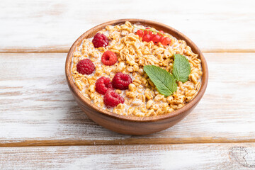 Fototapeta na wymiar Wheat flakes porridge with milk, raspberry and currant in wooden bowl on white wooden background. Side view.