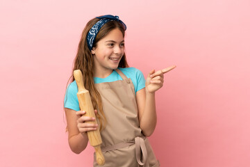 Little caucasian girl holding a rolling pin isolated on pink background pointing finger to the side and presenting a product