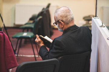Elderly man sitting on the chair at church service and reading from Bible.