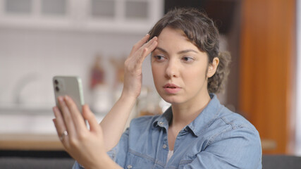 At home, young woman is using her mobile phone as a mirror and checking her skin. Beauty, skincare concept.