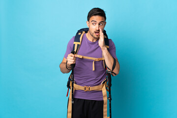 Young caucasian man with backpack and trekking poles isolated on blue background whispering something with surprise gesture while looking to the side