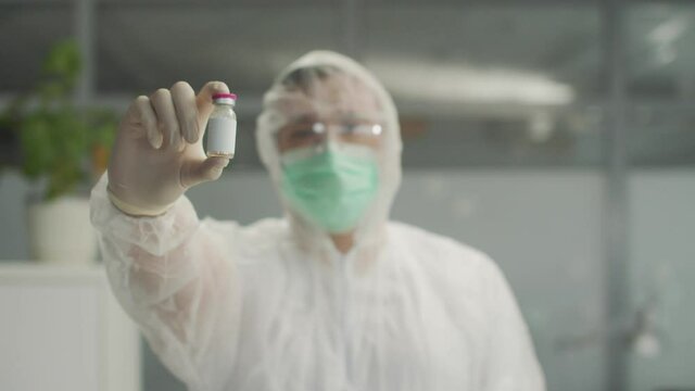 An unrecognizable doctor in a bacteriological protective suit holds a close-up vaccine in his hand and nods his head