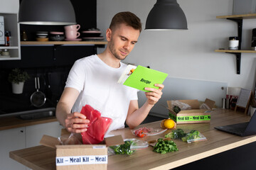 Happy Caucasian white man Unpack Online Home Food Delivery. Box with packed tuna, shrimp, vegetables and recipe card on a kitchen background. Food delivery services. Dinner Set from restaurant