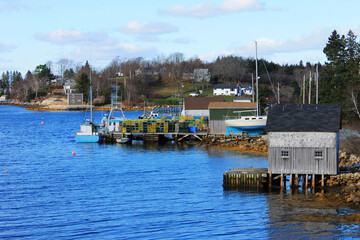 Fototapeta na wymiar View along the coastline of a Nova Scotia fishing village. Fish sheds, fishing boats, a sailboat on land, lobster traps stacked on a pier.