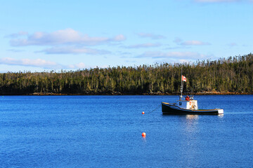 A small fishing boat anchored in a calm bay, Nova Scotia. Calm blue water, blue sky with light clouds, tree covered coastline.