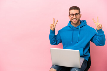 Young man sitting on a chair with laptop showing victory sign with both hands