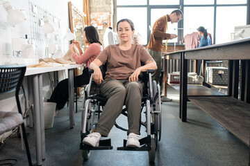 Fototapeta na wymiar Young disable female in casualwear sitting in wheelchair by workplace against group of colleagues