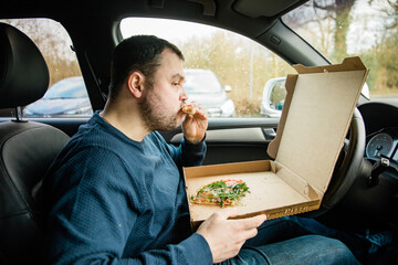 Man with beard eats pizza in his cart. Eat takeaway food in the car. Due to closed restaurants...