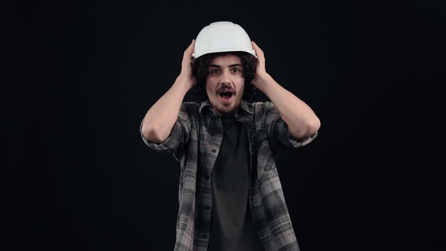 A surprised engineer wears a special helmet on his head, dressed in a plaid shirt, on a black background, in the studio, he says wow with his hands to his mouth, Industrial concept. People's emotions.