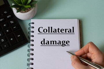 A woman's hand writes in a notebook the words COLLATERAL DAMAGE on the background of a calculator