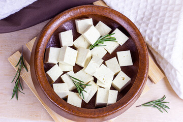 Top view of raw organic vegetarian tofu cubes in round bowl with fresh rosemary on wooden background.