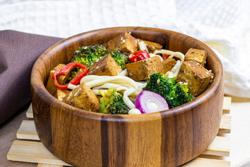 Homemade vegetarian fried tofu with udon noodles and vegetables (broccoli, pepper and onions) in the wooden bowl on light kitchen background.