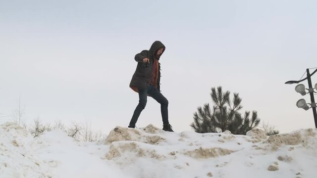 male photographer in black jacket and hood walks along the top of snowy mountain and is ready to take some photos. The naturalist photographer in his hands with camera goes forward and falls comically