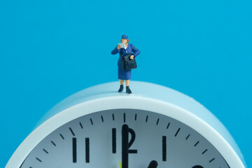 Miniature people toys conceptual photography. Delivery on time service. Postman courier standing above the clock, isolated on blue background.
