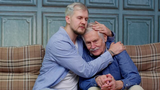 Young man asking for apologizes to offended stressed elderly senior father sitting together on couch