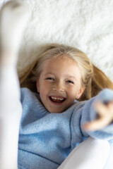 Cute little caucasian girl seven years old with blonde hair lying on bed at home in bedroom. Kid wearing stylish cozy knitted sweater blue color. Happy Child smiling