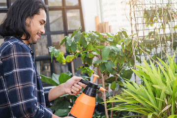 Smiling, happy young asian man, gardener is holding a water sprayer to water the plants with a spray in free time activities, sunshine in the morning at home,apartment.Lifestyle, hobby people concept.