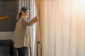 businesswoman traveller standing in hotel room by window with sunlight through curtain