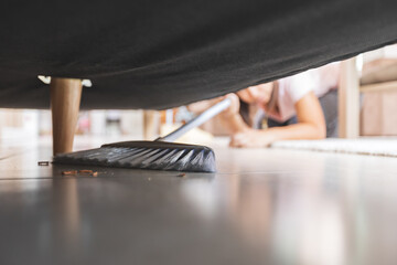 Asian woman cleaning and sweeping dust the floor under the sofa with a broom in the living room....