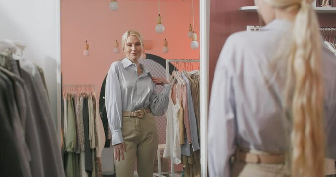 A Back View Shooting of a Blonde Woman Buyer Being in a Clothing Shop Standing in Front of The Mirror and Trying on a Jacket. Beautiful Cheerful Young Woman Choosing Clothes