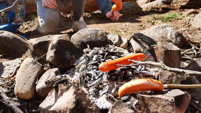 Sausages deliciously barbecued and moved around over open charcoal camp fire using carved wooden sticks. Sunny day covid-19 safe family hiking trip to the forest. VISIBLE HANDS AND FEET.