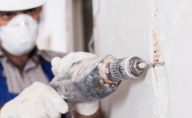 Male worker repairing construction with drill in mask and helmet.