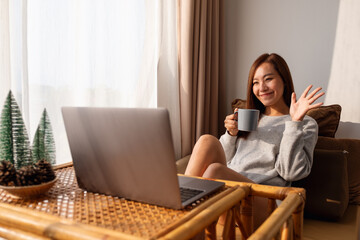 A beautiful young asian woman using laptop computer for video call while sitting and drinking coffee at home