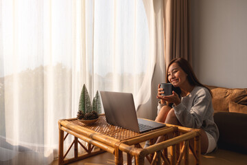 A beautiful young asian woman using laptop computer for video call while sitting and drinking coffee at home