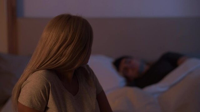 Couple issue. Night insomnia. Relationship problem. Anxious disturbed woman sitting on in bedroom with sleeping husband behind her.