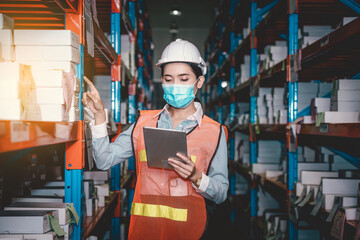 Industry manager and worker under checking about production at factory warehouse by tablet. They wearing safety uniform hard hat ,face mask to protect virus in new normal concept.