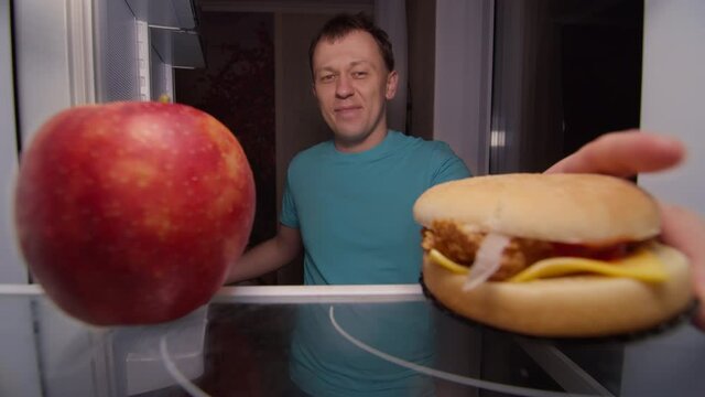 happy man opens the refrigerator and makes a choice between healthy and unhealthy food, apple or burger, unhealthy food in priority