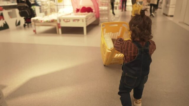 Cute One Year Old Toddler Pushing Yellow Shopping Basket Inside Ikea In Spain. following shot