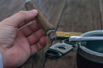A man's hand holds a smoldering cigar over a wooden table against the background of a guillotine, a...